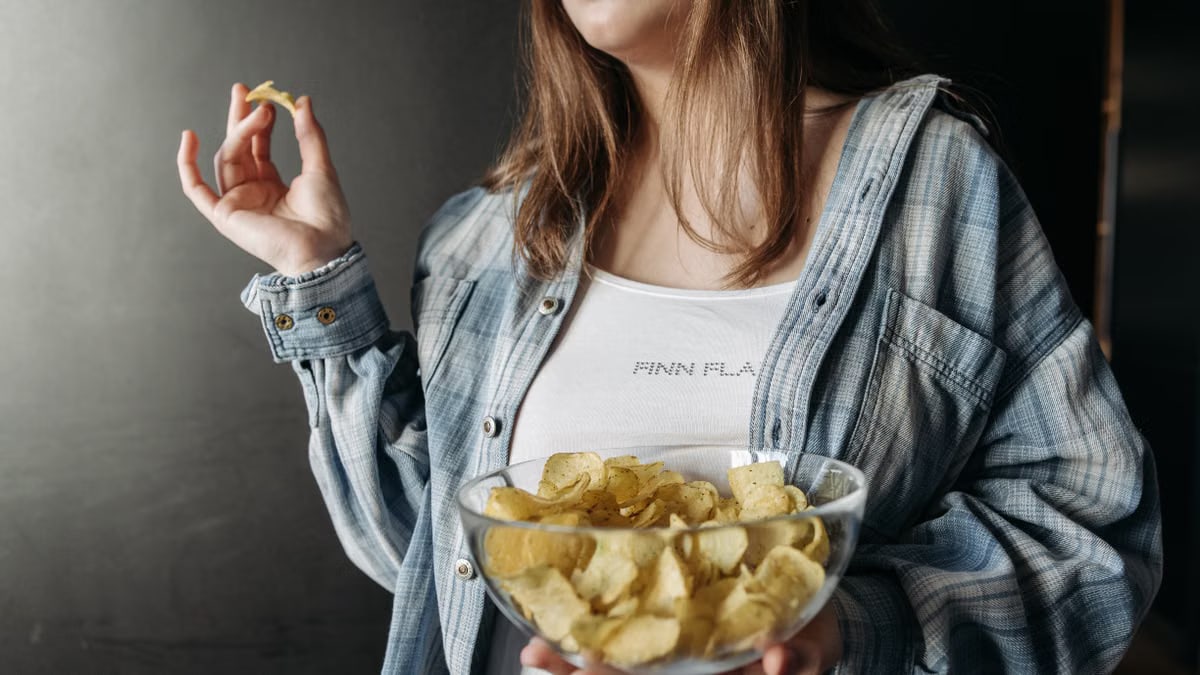 Woman in plaid shirt holding a bowl of potato chips indoors. Casual lifestyle image.